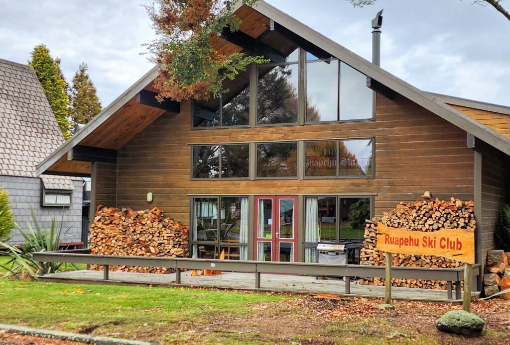Turoa Lodge, a two-storey timber and glass building among trees at Ohakune, with firewood stacked on the deck